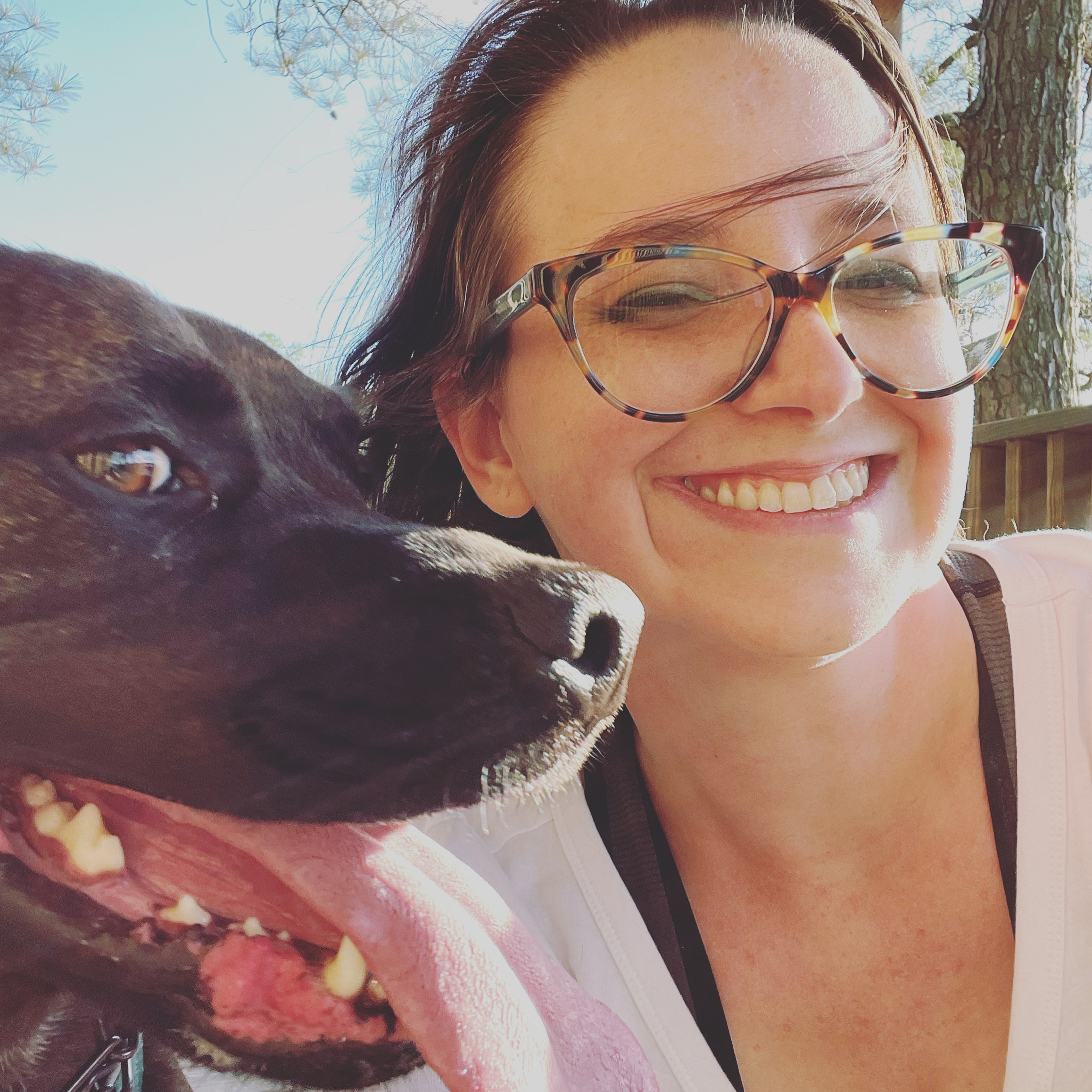 A picture of a young woman with glasses smiling happily with the cutest pupper in the world in the foreground.